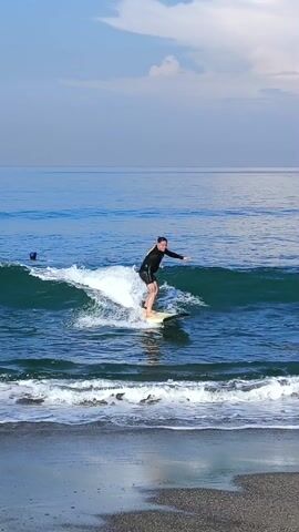 A man wearing a black wetsuit with yellow accents surfs towards the shore on a light-colored surfboard. He loses his balance near the beach, falling into the water. Other individuals are visible further out in the ocean, also on surfboards. The sky is partly cloudy, and the water is a deep blue. The beach appears to have dark sand.