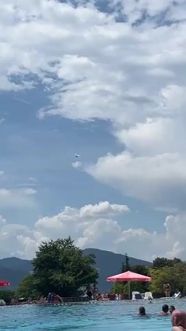 A hang glider is visible in the sky above a crowded outdoor swimming pool. The pool is filled with people swimming and relaxing. Trees and mountains are in the background under a partly cloudy sky. One person is seen entering the pool from the edge.