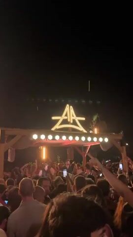 A nighttime scene shows a crowded outdoor venue, likely a bar or club. In the background, a wooden structure with a canopy is illuminated by strings of lights and spotlights. A large, stylized neon sign above the structure displays the letters "AA" in a unique font. A crowd of people is visible, many with their hands raised, dancing and enjoying the atmosphere. Some individuals are holding up their phones, possibly recording the event. The lighting is dim, with red and yellow hues dominating the scene, creating a vibrant and energetic ambiance. A palm tree is visible to the right of the structure, suggesting a tropical or warm climate location.