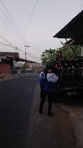 A group of people are gathered around a pickup truck loaded with bicycles. The truck is parked on a street next to a building with a covered porch and a white fence. A man in a blue shirt and white vest is seen handing a bicycle to someone in the truck. Another person in a blue shirt and white vest, and another person in a grey jacket are also visible. A man in a green jacket is sitting on the porch, looking at his phone. A red and white flag is hanging from the porch. The sky is overcast.