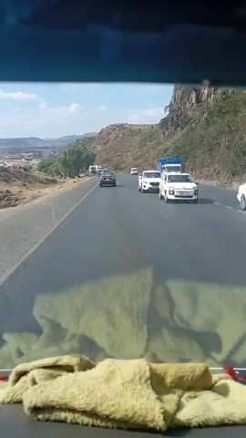 The video is taken from inside a moving vehicle, looking out through the windshield. The dashboard is visible at the bottom of the frame, covered with a light yellow, fuzzy cloth. The road ahead is paved and straight, with several vehicles in the distance, including a white van with "Radar" written on its side. To the left of the road is a relatively flat landscape with scattered trees and buildings. A large, rocky hill covered with vegetation rises up to the right of the road. The sky is blue with some scattered clouds, and the sun is shining.