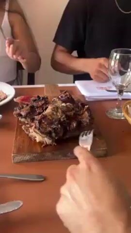 The video shows a group of people dining at a restaurant. A large piece of grilled meat on a wooden cutting board sits at the center of the table. A person wearing a silver watch with a brown leather strap cuts into the meat with a knife. A woman in a white tank top sits to the left of the meat. A person in a black t-shirt sits to the right. The camera then pans to show the view out the window. The restaurant appears to be located in a rural setting with trees, grass, and a horse-drawn carriage visible outside.