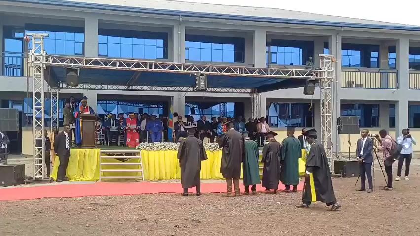 Here is a description of the scene in the video:
The scene appears to be a graduation ceremony taking place outdoors. A stage is set up in front of a modern building with large windows. On the stage, several people are seated, and one person is standing at a podium, presumably giving a speech. In front of the stage, a group of graduates dressed in academic robes and caps are standing. Several people are taking photographs or recording the event with cameras and phones. The ground appears to be unpaved. A red carpet leads to the stage. The weather seems overcast.