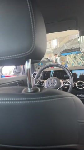The video shows the interior of a car, likely a Mercedes-Benz, seen from the back seat. The camera is focused on the back of the headrests and the steering wheel in the front. In the background, through the car's windows and possibly a showroom or garage door, other vehicles can be seen, including one on a lift covered with a tarp. A person in a white shirt is visible near the lifted car. The overall impression is a car dealership or repair shop environment.