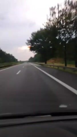 The video shows a view from inside a vehicle moving along a highway. The highway has multiple lanes and is bordered by a guardrail and trees. In the distance, several wind turbines are visible against a cloudy sky. The sun appears to be setting or rising, casting a dim light. The camera is mounted inside the car, looking forward, and the dashboard is partially visible at the bottom of the frame. There are no people visible in the video. The road is empty except for a few distant vehicles. The overall scene depicts a journey on a highway during twilight.