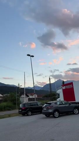 This video captures a serene sunset scene in Völs, Austria. The sky is painted with soft hues of pink and orange clouds against a tranquil blue backdrop. In the foreground, two cars are parked on an asphalt surface, a black SUV and a gray crossover. Behind them, a white semi-truck with a red side bearing the text "MPREIS" is visible.

Further in the background, a charming Austrian village unfolds with houses featuring red-tiled roofs nestled amongst lush green trees. Power lines crisscross the landscape, supported by tall utility poles, hinting at the infrastructure of a modern yet nature-embracing setting. Beyond the village, majestic mountains rise, their peaks softened by the twilight. The overall atmosphere is peaceful, suggesting a calm evening in a picturesque European locale. The time of day is clearly sunset, with the soft, warm light casting a gentle glow over the scene. The weather appears to be clear, with only scattered clouds adorning the sky.