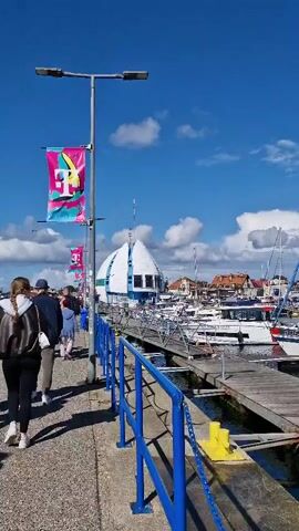 The video shows a sunny day at a harbor. A group of people are walking along a paved walkway beside a blue railing. In the background, several sailboats and yachts are docked in the water. There is a unique, white building with a pointed roof that resembles a spaceship, and other buildings with red roofs are visible further in the distance. Banners with a pink and teal design featuring the letter T are hanging from lampposts.