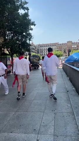 The video shows a group of people walking down a paved city street. Most of them are wearing white shirts and light-colored shorts or pants, with red scarves around their necks. They appear to be walking away from the camera, towards a truck in the distance. The street is lined with buildings, trees, and a metal railing. There are some shops and a stage or platform in the background. The sky is clear and the sun is shining. Some people are also seen on the other side of the street, and in the background, there are more buildings and trees. The overall atmosphere appears to be a daytime event or festival.