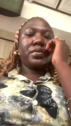 A dark-skinned woman with light brown braided hair and a patterned headband is captured in a chest-up shot, looking directly at the camera. She wears a light green and dark green patterned button-up shirt, gold hoop earrings, and multiple rings on her right hand. Her right hand is positioned near her chin and mouth. A silver bracelet is visible on her left wrist. The background, slightly out of focus, shows a wall, a television screen in the upper left, and a white security camera in the upper right. Ambient background noise is audible.