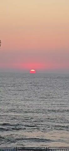 A video clip shows a sunset over the ocean. The sun is a bright red orb partially submerged below the horizon, casting a vibrant pink glow across the sky and the rippling water. The scene is serene and peaceful, with the gentle waves creating a subtle texture on the ocean's surface. The sky transitions from a soft pink near the horizon to a lighter hue as it ascends. The sun appears to be slowly setting, with its position in the frame remaining relatively consistent. The overall atmosphere is one of natural beauty and tranquility.