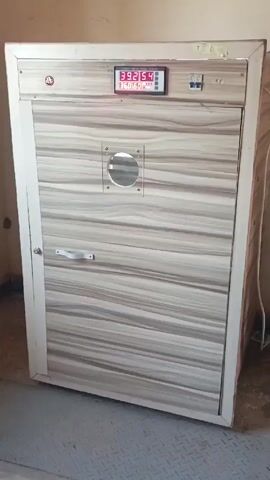 The video shows a freestanding, wooden-cabinet-style egg incubator. The incubator's door is a light gray-brown striped wood-look pattern, and the frame is white. A digital display on the top of the incubator shows readings in red numbers. Below the display are two toggle switches. A small, round window is in the upper part of the door. The door has a white handle on it. The incubator is in a room with beige walls and a gray metal floor.  A white electrical cord is hanging from the side of the incubator.