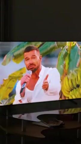 The video shows a man singing into a microphone. He has short brown hair, a beard, and is wearing a white shirt. The background behind him appears to be foliage and bananas. The scene is being displayed on a television screen with a reflective black base. A small dish with items inside is visible on the stand below the television.