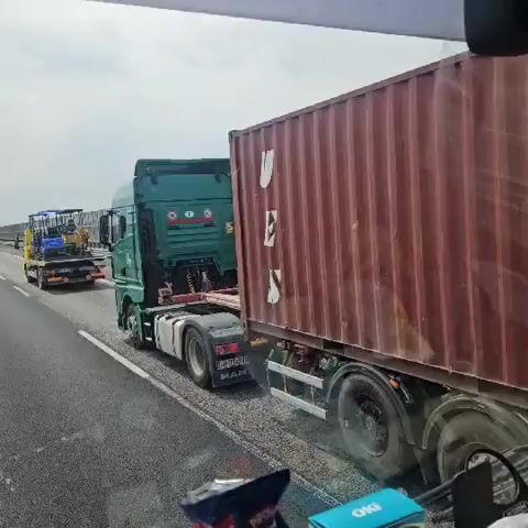The video shows a convoy of trucks on a highway. The camera is inside a vehicle, likely a truck or bus, and the view is through the windshield. In the foreground, the back of a green semi-trailer truck is visible, towing a large red shipping container with the letters "UES" printed on its side. Behind this truck, another truck is visible, this one yellow, carrying two small excavators. The excavators are blue and yellow. The road appears to be a multi-lane highway with a barrier on the right side, likely a sound barrier. The sky is overcast. The trucks are traveling in the same direction.