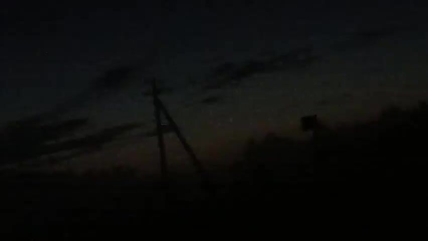 The video shows a dark outdoor scene at dusk or night. A lone utility pole with a crossarm is visible in the center-left of the frame. To the right of the pole, there is a dark silhouette of trees and a possible signpost. The sky is dark with some faint clouds visible, and there appears to be some light pollution or stars. The overall impression is of a rural or undeveloped area.