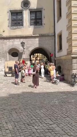 A group of people are gathered in a courtyard paved with cobblestones. In the background, there is an archway leading to a building. Several people are holding bouquets of flowers and one person appears to be throwing something into the air, creating a cloud of small particles. Some people are dressed casually, while others are wearing more formal attire, including one person in a white suit and another in a brown dress. There are also some red balloons visible. The environment appears to be outdoors on a sunny day, with shadows cast by the buildings. A small dog is also visible on the right side of the frame.