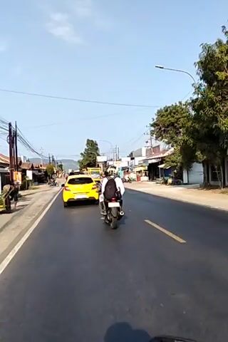 The video is taken from the perspective of someone riding a motorcycle. The rider is on a paved road with a yellow car in front of them, also moving forward. To the right of the road, there are several buildings, some appearing to be shops or businesses. Trees and utility poles with wires are visible along the sides of the road. The sky is clear and blue, indicating a sunny day. There are other vehicles on the road, including a yellow truck. The rider is wearing a backpack. The overall scene depicts a typical road with traffic in what appears to be a somewhat rural or suburban setting.