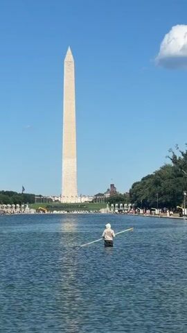 The video shows the Washington Monument and the Reflecting Pool on a clear day. A person is standing in the shallow water of the Reflecting Pool, holding a long pole. The person is wearing a white hat and a light-colored shirt. The Washington Monument is tall and white, with a pointed top. Trees line the edge of the Reflecting Pool, and buildings can be seen in the background behind the monument. The sky is blue with a few white clouds. The water in the Reflecting Pool is calm and reflects the sky and the surroundings. The person in the water appears to be cleaning or maintaining the pool.