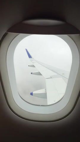The video shows a view from an airplane window. The wing of the airplane is visible, extending out into a hazy sky. The wing is white with blue accents at the tips of its flaps. The window frame is gray and oval-shaped. The sky outside appears overcast and white, with some darker cloud formations visible below the wing. The overall impression is of an airplane in flight.