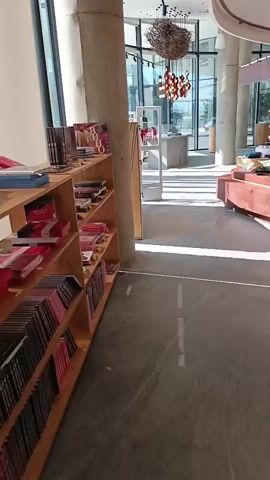 The video pans across the interior of a well-lit store. The store features a curved wooden display counter with multiple tiers, showcasing various products, including what appear to be bottles and boxes. Behind the counter, floor-to-ceiling shelving units are stocked with more items, including many bottles that resemble wine or spirits. To the left of the frame, a wooden bookshelf is filled with colorful items, possibly books or merchandise. Large windows in the background allow natural light to enter, illuminating the space. The flooring is a polished gray material with subtle veining. The overall ambiance suggests a high-end retail environment, perhaps a boutique wine shop or a specialty food store. There are no people visible in the video.