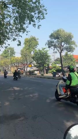This is a vertical, outdoor video filmed from a motorcycle, showing a street scene on a sunny day with clear blue skies. The camera perspective is from behind the handlebars, and the motorcycle's side mirror is visible in the bottom right corner, reflecting the road and other vehicles.

The road is paved and appears to be a multi-lane street with a clear white line marking the edge of the lane. There is a paved sidewalk on the right side of the road, separated by a curb.

On the road, several motorbikes and at least one black SUV are visible, all traveling in the same direction as the camera. One motorbike in the foreground on the left has a rider wearing a green jacket and a helmet. Another motorbike in the middle of the road has a rider wearing a dark helmet and a backpack. Further down the road, more motorbikes are seen. A black SUV is positioned to the right of the road, slightly ahead of the motorbikes.

Lush green trees line the left side of the road, with their branches extending over the street, creating dappled shadows on the asphalt. On the right side of the road, beyond the sidewalk, there are buildings, some with red-tiled roofs, and more trees. In the distance, the scene suggests a suburban or semi-urban environment. The overall atmosphere is one of everyday traffic and street life under good weather conditions.