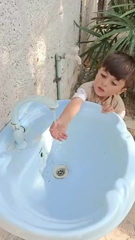 A young boy, seen from about the chest up, reaches into a blue outdoor sink where water is running from the tap. The sink is situated outside, next to a light-colored wall with some green piping, and a green palm-like plant is visible in the background. The boy, with brown hair and a tan shirt, looks at the water with interest as he interacts with it.