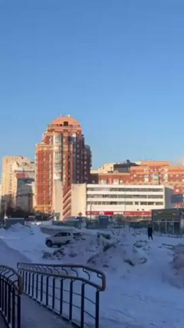 The scene depicts a winter landscape in an urban setting. Snow covers the ground, with drifts along a pathway. In the background, several multi-story buildings are visible, a tall brick building with a rounded top being the most prominent. A car is parked amidst the snow. A lone individual is walking away from the camera on the sidewalk in the distance. The sky is clear and blue.