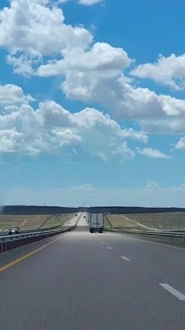 A truck drives on a highway under a blue sky with white clouds. The highway has multiple lanes and a yellow line marking the edge. The landscape on either side of the highway is flat and appears to be dry land with some sparse vegetation. There are guardrails along the side of the road.