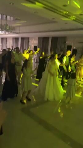 The video shows a wedding reception with a group of people dancing. The bride, in a white wedding dress, is in the center of the frame, surrounded by guests. The room is brightly lit with green spotlights. A DJ booth with speakers is visible in the background. Several guests are wearing formal attire, including women in dresses and men in suits. Some children are also present and dancing. The atmosphere appears festive and celebratory.