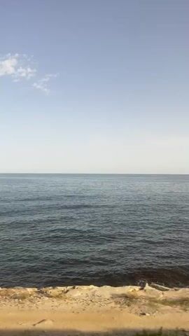 The video shows a view of the ocean from a moving vehicle. The camera is positioned to capture the horizon line, which is mostly clear with a few wispy clouds in the sky. The water is a deep blue-green with small waves rolling towards the shore. The foreground of the video shows a rocky embankment with some sparse vegetation, suggesting a coastal road or path. The movement of the vehicle causes the scene to shift slightly, but the overall view of the ocean remains consistent. There are no people or specific objects visible in the video other than the natural elements of the sea and shore. The lighting suggests it is daytime, likely late afternoon or early morning due to the soft light and clear sky.