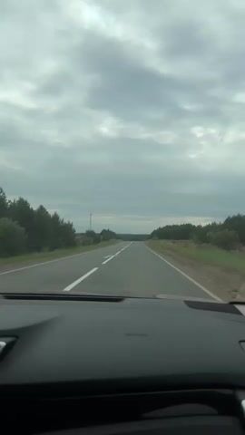The video shows a POV shot from inside a car driving on a paved road under a cloudy sky. The road is bordered by trees on both sides. There are white lane markings on the road. A tall, slender tower-like structure is visible on the left side of the road in the distance. The view ahead shows a long stretch of road disappearing into the horizon. The interior of the car, specifically the dashboard, is partially visible at the bottom of the frame. The overall atmosphere is overcast and suggests a journey.
