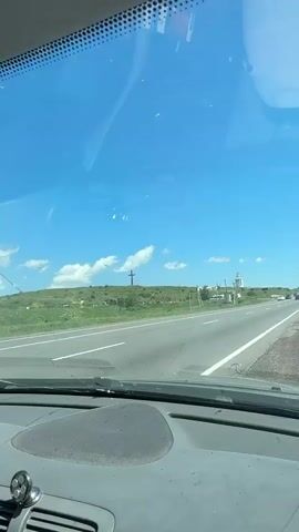 The video captures a view from inside a vehicle traveling on a road. The first frame shows a white minivan driving ahead on the right side of the road. The sky is blue with scattered clouds. The second, third, and fourth frames show a large, dark cross on top of a green hill. The cross appears to be made of metal and is quite tall. There are some buildings and other structures near the base of the hill, and some trees and bushes are visible. In the foreground of the first frame, a portion of the vehicle's dashboard and windshield is visible. The overall environment suggests a rural or semi-rural setting with a clear, sunny day.