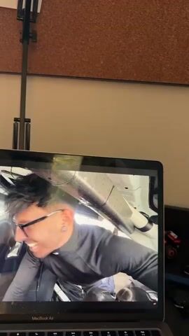 The video captures a close-up shot of a laptop screen displaying a man in what appears to be a racing environment. The man, with dark hair, glasses, and an ear piercing, is smiling and looking towards the camera. He is wearing a dark top, possibly a racing suit, and a white helmet with a black and white checkered flag emblem is visible on his head. The interior of the vehicle he is in shows parts of a roll cage and other racing equipment.

Visible text on the screen includes subtitles: "...say some prize soon, we'll see where we are. See if we can actually compete in that. So now it's time to get for me." The laptop itself is a "MacBook Air".

In the background and foreground of the shot, a cork board is visible, along with a black metal stand, possibly for a light or microphone, which extends upwards. There are also some faint, illegible writings on papers attached to the cork board. A small red object, possibly a toy car or miniature, is on the desk next to the laptop. The setting appears to be an indoor space, possibly a home office, in Novate Milanese, Italy. The lighting suggests an indoor environment.