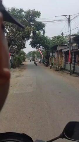 The video shows a street view from the perspective of someone on the back of a motorcycle. The road is paved and appears to be in a rural or suburban area, lined with houses and trees. Several people are riding motorcycles in the same direction, some wearing helmets and others not. There are also some parked motorcycles and bicycles visible. In the background, there are red and white flags, possibly indicating a celebration or event. The sky is overcast. The person filming is looking forward, and their face is partially visible in the foreground.