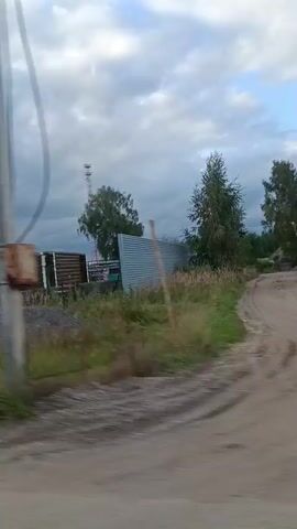 The video shows a view from a moving vehicle, likely a car, passing by a rural or semi-industrial area. The landscape features vegetation, including tall grass and trees. Several structures are visible, including what appears to be a telecommunications tower, some wooden buildings, metal sheds, and stacks of construction materials like pipes and flat panels. One building has a red roof and a sign with Cyrillic text. The sky is partly cloudy with a mix of blue sky and grey clouds. The road surface in the foreground is dirt or gravel. The overall impression is of a utilitarian or industrial area on the outskirts of a town or village.