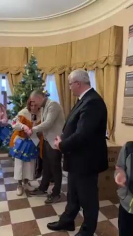 The video captures a scene indoors during what seems like a festive event. The floor has a checkered pattern of dark brown and light brown squares. To the left, there's a Christmas tree decorated with ornaments and lights. Several individuals are present.

A man in a dark suit is handing out blue gift bags with cartoon-like designs. He gives a bag to a younger woman with long hair who's wearing a white t-shirt, jeans, and a pink plaid jacket. Another woman stands nearby, dressed in a traditional Ukrainian outfit. A man in a light-colored jacket is also present, seemingly involved in the gift distribution. 

Behind them, there are gold curtains and a wall with some kind of exhibit. To the right of the man in the suit, there's an LG branded cardboard box. Another person stands to the right of the box. The atmosphere appears to be celebratory, and there's some chatter and applause in the background.