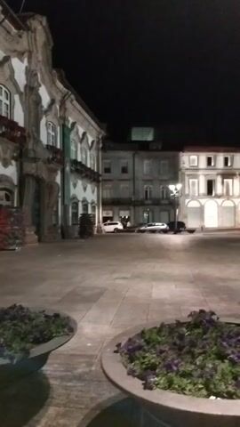 The video shows a nighttime view of an outdoor public space, likely a plaza, with cobblestone paving. In the foreground, two large, round planters filled with purple flowers are visible. In the midground, several multi-story buildings line the plaza. Some of these buildings have illuminated windows and arched doorways. A streetlamp stands prominently in the foreground, slightly off-center. Several cars are parked or driving in the distance. A few people can be seen walking in the background on the right side of the frame. The overall atmosphere is calm and quiet.