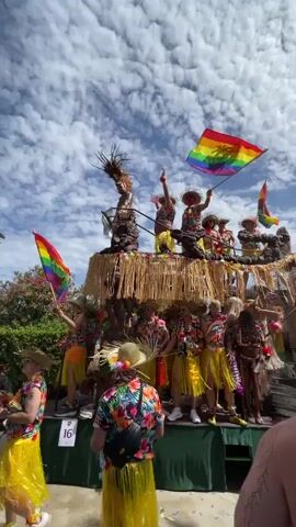 The video shows a festive parade float decorated with a Hawaiian theme. The float is adorned with straw and tropical elements, and features a group of people dressed in colorful, floral shirts and yellow grass skirts. Some individuals are wearing straw hats and leis. Two rainbow flags are prominently displayed and being waved. The people on the float appear to be dancing and celebrating. The background consists of a bright blue sky with fluffy white clouds and some greenery from trees. A number "16" is visible on a sign on the lower part of the float.