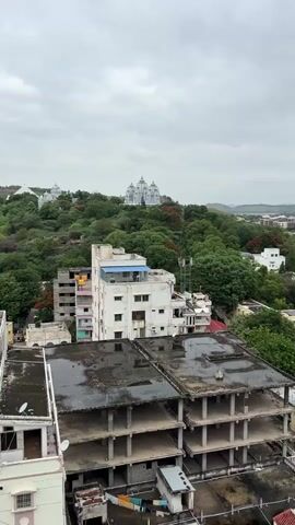 The video captures a panoramic view of a city from an elevated perspective. In the foreground, unfinished buildings with exposed concrete structures and flat, dark roofs dominate the scene. The roofs appear wet, suggesting recent rain. In the midground, several multi-story buildings are visible, some painted white and others in various colors. One white building in the center has a blue rooftop structure. Green trees are abundant throughout the city, with some trees bearing red flowers. In the background, a large, white, ornate building, possibly a temple or a palace, sits atop a hill covered in lush green foliage. Further in the distance, rolling hills and a hazy sky with scattered clouds complete the landscape. The overall impression is of an urban environment nestled within a verdant, hilly region under an overcast sky.