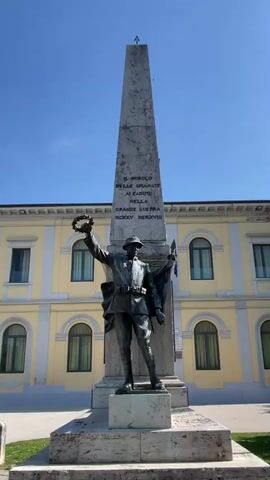 A low-angle, full-shot video shows a bronze statue of a soldier holding a wreath and a flag, standing in front of a tall obelisk inscribed with text in Italian. The statue and obelisk are situated in front of a pale yellow building with arched windows. The sky above is clear and blue. The statue is dressed in military attire, including a helmet, uniform, and belt. The soldier's left arm is raised, holding a laurel wreath. His right arm is bent, holding a flag. The obelisk is weathered and appears to be made of stone. The inscription on the obelisk reads "IL POPOLO DELLE SPIANATE AI CADUTI NELLA GRANDE GUERRA MCMXV-MCMXVIII". The building in the background is a large, multi-story structure with a symmetrical facade. The overall scene suggests a war memorial.