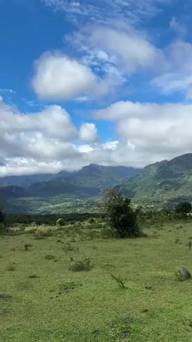 The video shows a landscape featuring a green field in the foreground, transitioning into mountainous terrain in the background. The sky, initially blue with scattered white clouds, gradually becomes overcast as the video progresses. The mountains in the distance are covered with lush green vegetation, and some peaks are obscured by low-hanging clouds. There is a lone tree and some smaller shrubs in the field. The camera slowly pans from right to left, offering a broad view of the scenery.