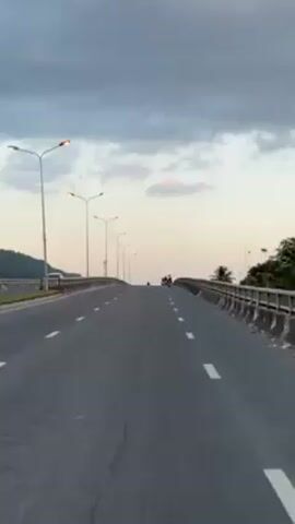 A view from the middle of a highway bridge looking uphill at dusk. A pair of motorcycles is visible ahead driving in the same direction, with mountains visible in the background. The highway is mostly empty, with lane markers clearly visible on both sides of the bridge. Evenly spaced street lamps line the bridge's edge on both sides. The sky is a mix of light blue and gray clouds.