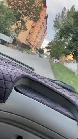 A dashcam view from inside a vehicle shows a street with a tall, tan apartment building on the left and trees and grass on the right. A dark-colored car is visible in the distance driving away from the camera. The vehicle appears to be moving forward.