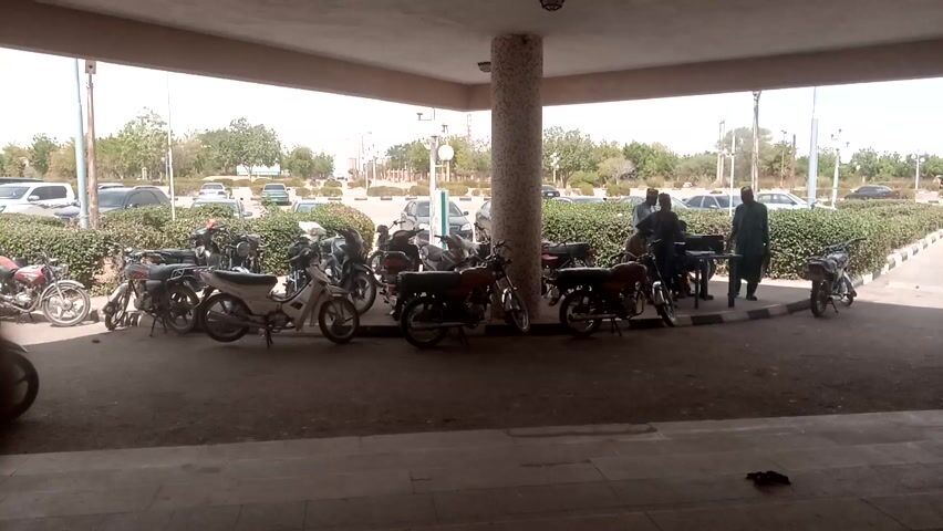 The video displays an outdoor scene under a building's concrete overhang, which provides shade. The ground is paved, and there are steps leading down in the immediate foreground.

Numerous motorcycles of various models and colors are parked in rows under the overhang, occupying most of the frame. A light-colored motorcycle is prominently parked on the left side in the foreground.

Several men are present in the scene. On the right side, two men, both wearing long tunics (one dark, one light-colored), are standing near a small, dark table, appearing to be interacting or conducting a transaction. Another man, also in a light-colored long tunic, walks from right to left in the background. On the far left, a man in a light-colored long tunic leans against a large, mosaic-tiled pillar that supports the overhead structure. A person is partially visible in the bottom-left corner, sitting or crouching.

Beyond the shaded area, a sunny outdoor parking lot is visible, partially obscured by a line of green bushes. Several cars, including white SUVs and sedans, are parked in this area. In the far background, trees and some distant buildings can be seen under a bright sky. A brown market-style umbrella is also visible in the background, near the parked cars. The setting suggests a public space, possibly a motorcycle parking zone or a taxi stand, during daytime.