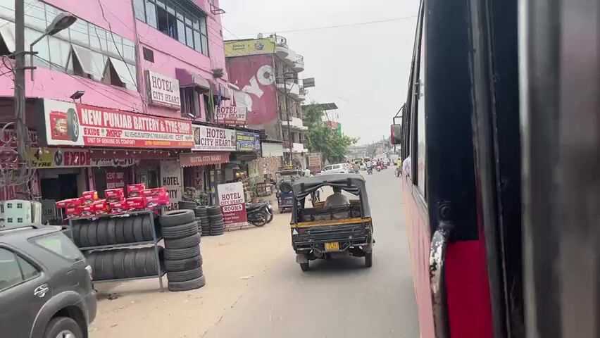 The video captures a street scene in Rupnagar, India, from the perspective of a moving vehicle, likely a bus. The camera pans across a bustling street lined with various businesses. Prominently displayed are signs for "HOTEL CITY HEART ROOMS ROOMS ROOMS" and "PUNJAB BATTERY & TYRE SHOP." Further down, another hotel sign, "HOTEL CITY HEART," is visible, along with a display of stacked tires. Other businesses include shops selling goods like snacks and what appears to be an automotive service or parts store, indicated by the MRF branding.

The street is active with traffic. Several auto-rickshaws, a common mode of transport in India, are present. One auto-rickshaw is directly in front of the camera vehicle, while another is further down the road. A few motorcycles are also seen, with people riding them. There are also cars and bicycles on the street, suggesting a mix of transportation methods.

People are visible in the scene, some operating vehicles, others walking or standing near the shops. The overall atmosphere appears to be that of a busy commercial street during daytime. The sky is overcast, indicating a cloudy day. The architecture of the buildings suggests a mix of older and newer constructions. Red and pink are dominant colors in the facade of the buildings on the left. One building features a large red OYO logo.

There is visible text on various signs. Besides the hotel and shop names, there are also smaller signs advertising services and products. The weather appears to be warm and humid, typical for the region. The overall impression is of a typical day in a moderately sized Indian town.