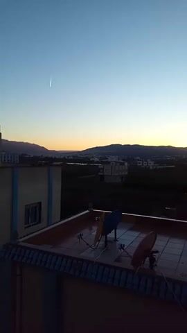 The video opens with a wide shot from a rooftop at what appears to be either sunrise or sunset. The sky displays a beautiful gradient of deep blue at the top fading into light blue and then warm orange and yellow hues closer to the horizon. A prominent, long, thin white streak, likely a contrail from an aircraft, is visible in the upper left portion of the blue sky.

In the foreground, on the rooftop, two satellite dishes are clearly visible: one blue and one red-brown, both pointing upwards. Below the rooftop, parts of other buildings with blue and yellow/beige walls are seen. In the mid-ground, there's a sprawling landscape featuring more buildings, some isolated and some clustered, interspersed with green fields. A distant mountain range outlines the horizon where the sun's light is most intense. The audio includes faint singing, possibly religious, followed by the sound of wind.

At 0:10, the camera begins to pan slowly to the right. The view shifts, revealing more of the surrounding urban or suburban area. Several white and beige multi-story buildings with various architectural details are visible. A road with some vehicles can be glimpsed between buildings and green areas. The sky maintains its gentle color gradient.

As the camera continues to pan towards the right (around 0:26), the landscape evolves to show a wider expanse of green fields and more buildings, including some with red-framed windows. Eventually, a distinct blue line appears on the distant horizon, suggesting a large body of water, likely the sea or ocean. The camera stops panning around 0:42, showcasing this coastal view under the soft, fading light of dawn or dusk. Throughout the pan, occasional faint sounds of traffic or distant activity can be heard.