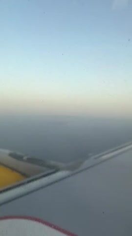 An aerial view from an airplane window shows a hazy sky fading into the horizon above a body of water. The wing of the plane is visible in the foreground, with a section of the engine on the left. A small, indistinct landmass or island is visible in the distance, partially obscured by atmospheric haze. The lighting suggests it is either dawn or dusk.