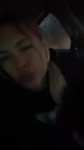 The video shows a woman in a dark vehicle at night. The environment is dark, but an outdoor light is visible in the background through a window with rain. The woman has blonde hair pulled back in a ponytail. She is wearing a dark hoodie. She looks at the camera, pouts her lips, and slightly smiles.