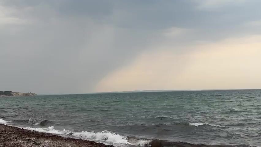 A wide shot of a choppy, greenish-blue ocean with waves breaking on a rocky shore with seaweed. The sky is overcast and grey, with a lighter patch of sky visible on the right horizon, possibly indicating a break in the clouds or a distant storm. A faint landmass can be seen on the far left horizon. The perspective is from the shore looking out to sea.