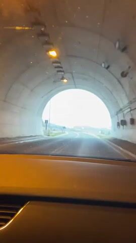 The video is filmed from the interior of a vehicle, looking out through the windshield. It begins with the vehicle inside a concrete tunnel, heading towards a bright, circular exit. An orange-yellow light fixture is visible on the tunnel's ceiling directly above the windshield.

As the vehicle exits the tunnel, the scene transitions to a two-lane asphalt road under a heavily overcast, gray sky. The landscape on either side of the road is rugged and somewhat barren. To the left, a rocky hillside with sparse, dry vegetation runs alongside the road. Several tall, modern streetlights are positioned at intervals along this side. To the right, the terrain is more open, featuring dry ground with scattered small trees and shrubs. In the distance, hills and mountains are visible, their peaks blending into the cloudy sky, some showing faint patches of lighter color, possibly snow or lighter vegetation.

Two road signs are visible on the left and right sides of the road. The left sign is triangular and black, while the right sign is round, white with a red border, and appears to depict two cars, suggesting a regulatory warning. The road ahead gently curves and extends into the distance. Music is audible in the background throughout the video. The setting appears to be a rural or semi-rural area in Provalija, Montenegro, based on the provided context.