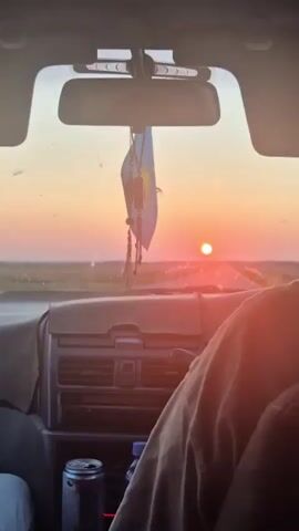 The video shows a view from inside a car looking out the windshield at a sunset. The sun is a bright orange orb low on the horizon, casting a warm glow over a flat, open landscape. A small flag of Kazakhstan is hanging from the rearview mirror. The dashboard of the car is visible in the foreground, with a person's arm resting on the center console. The overall impression is one of a peaceful journey during a beautiful sunset.